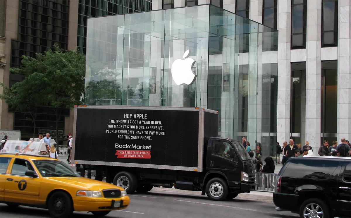 A black box truck parked outside a glass Apple store. Its side reads “Hey Apple. The iPhone 17 got a year older. You made it $100 more expensive. People shouldn’t have to pay more for the same phone.” above the Back Market logo