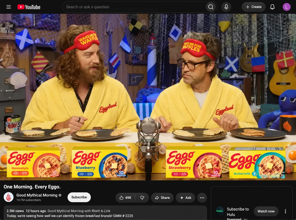 YouTube video titled “One Morning. Every Eggo.”: two hosts in yellow Eggoland robes and “Wake me for waffles” sleep masks tasting waffles